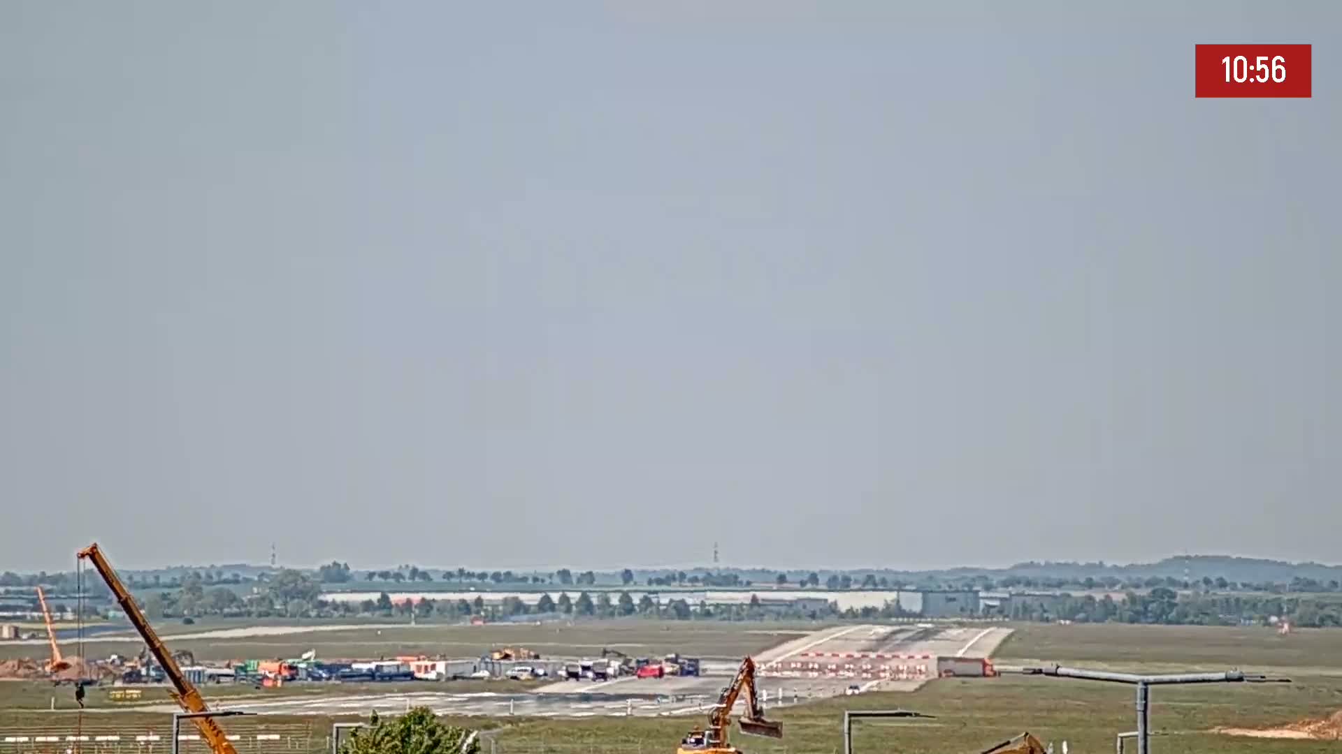 Under a hazy sky, construction equipment is visible on an airport runway surrounded by flat, green land.