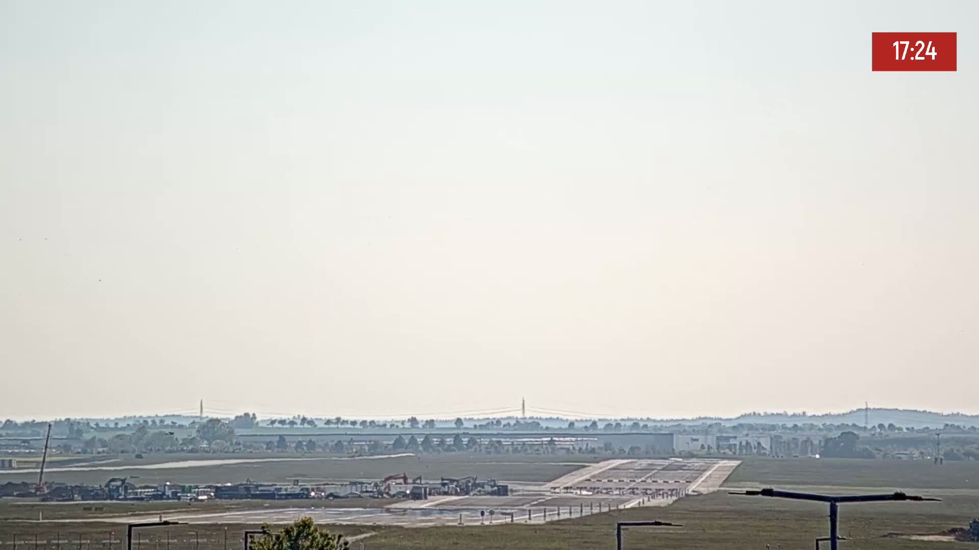 A hazy, sunny day shows construction equipment on an airport runway with a distant treeline in the background.