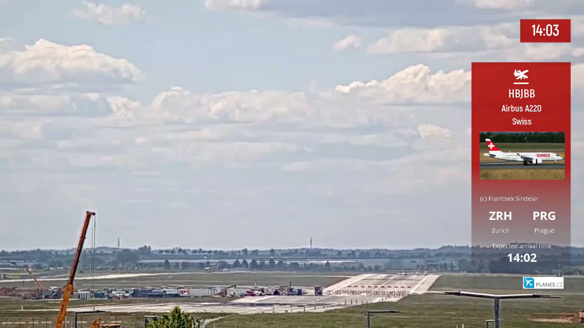A long shot of an airport runway under a partly cloudy sky shows construction equipment and a plane landing in the distance.