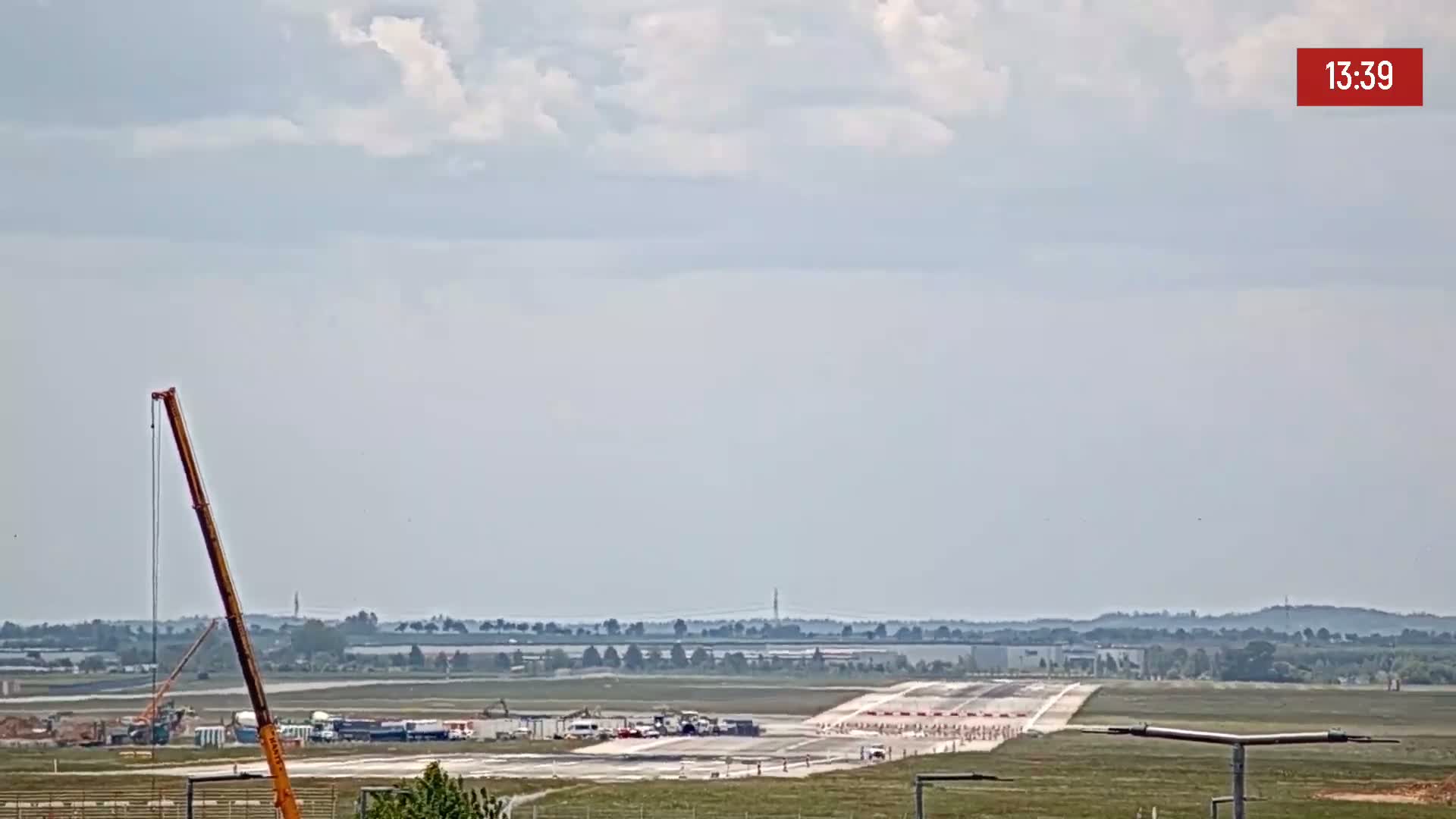 Under a partly cloudy sky, a long runway is visible with construction equipment and vehicles present near it.