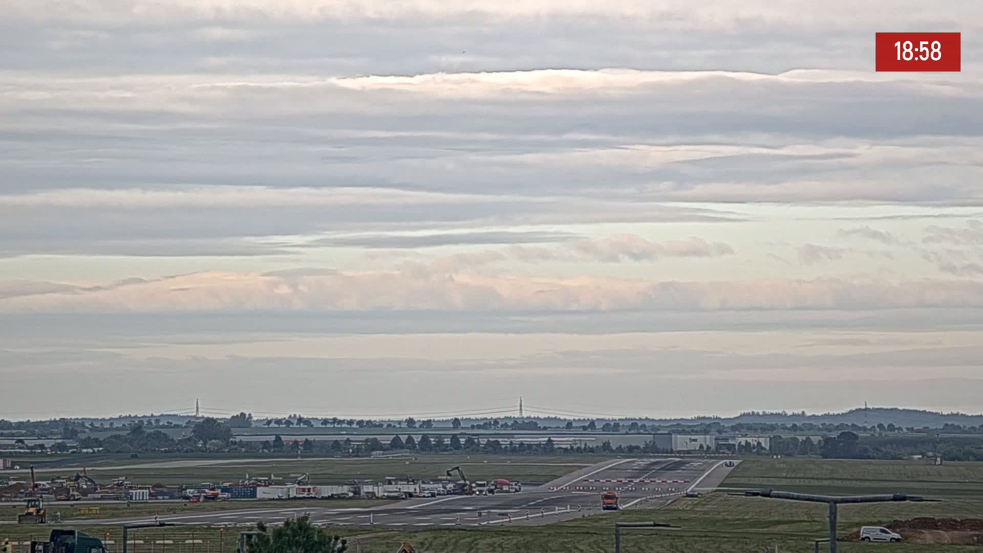 Under a mostly cloudy sky, an airport runway is shown with construction equipment and vehicles near the tarmac.