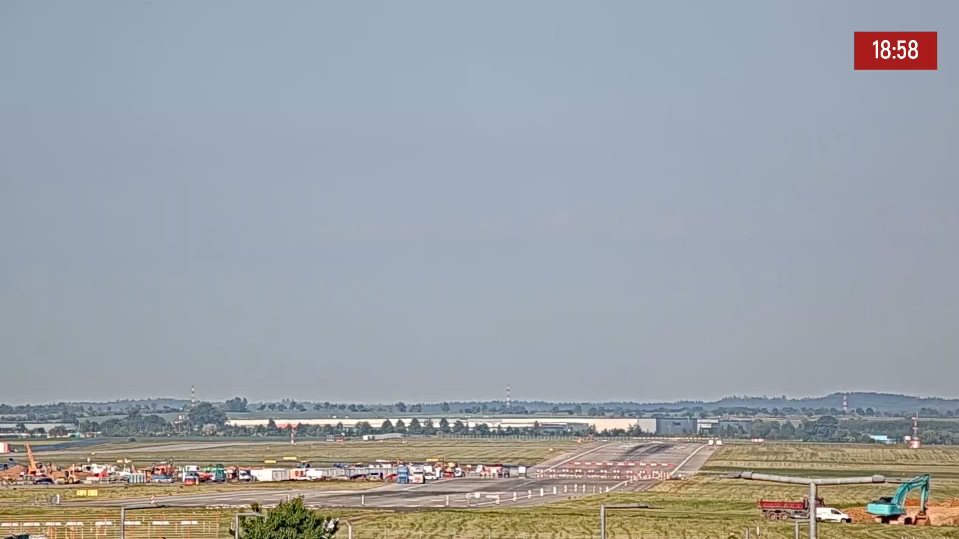 A long runway at an airport with construction equipment and vehicles is visible under a hazy, light-grey sky.