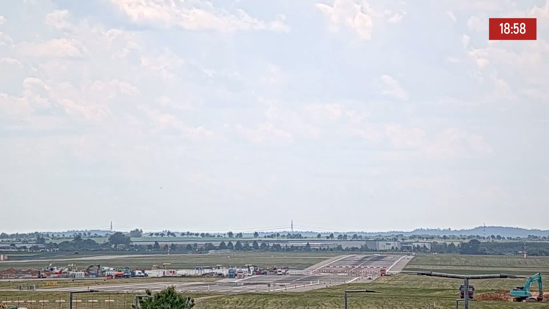 A long runway at an airport is visible under a partly cloudy sky, with construction equipment and vehicles parked nearby.