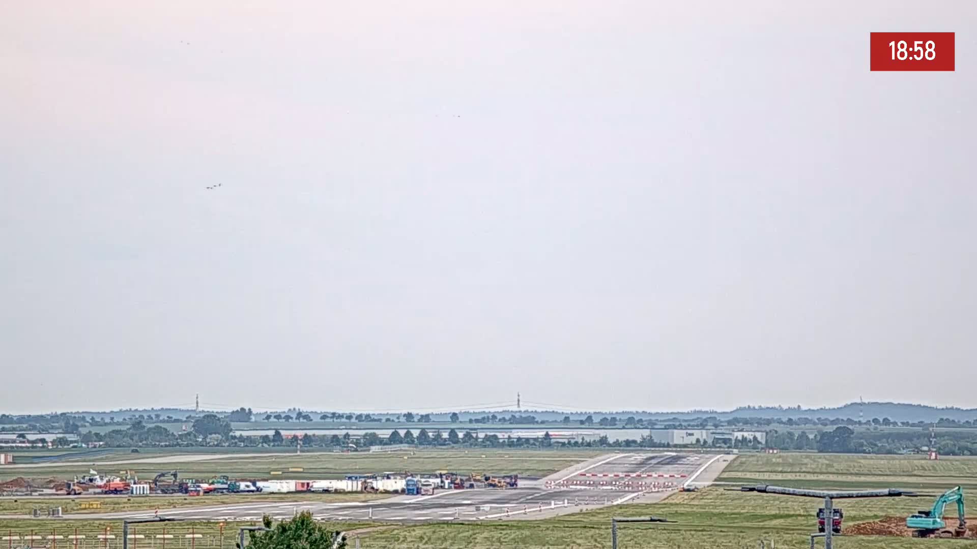 A long view of an airport runway under a hazy sky shows construction equipment and vehicles parked near the runway.