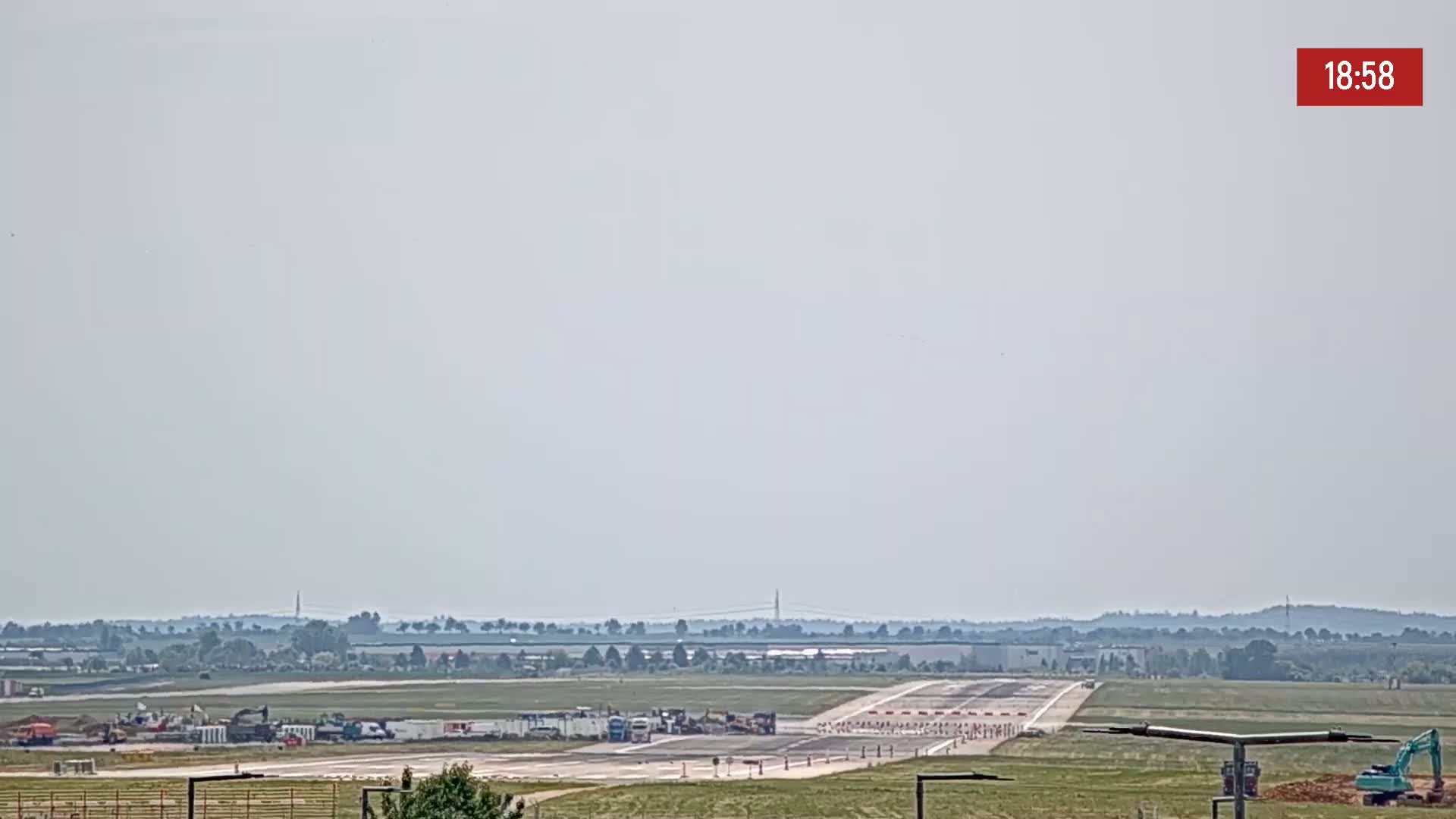 Under hazy skies, a long runway at an airport is shown with construction equipment and vehicles near the edge.