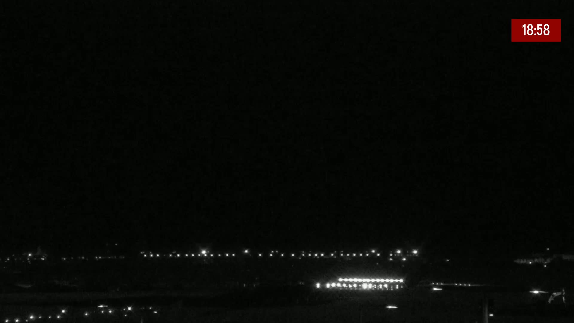 A nighttime, long shot shows a dark landscape with a line of distant lights and a brighter area of lights in the midground under a clear, dark sky.
