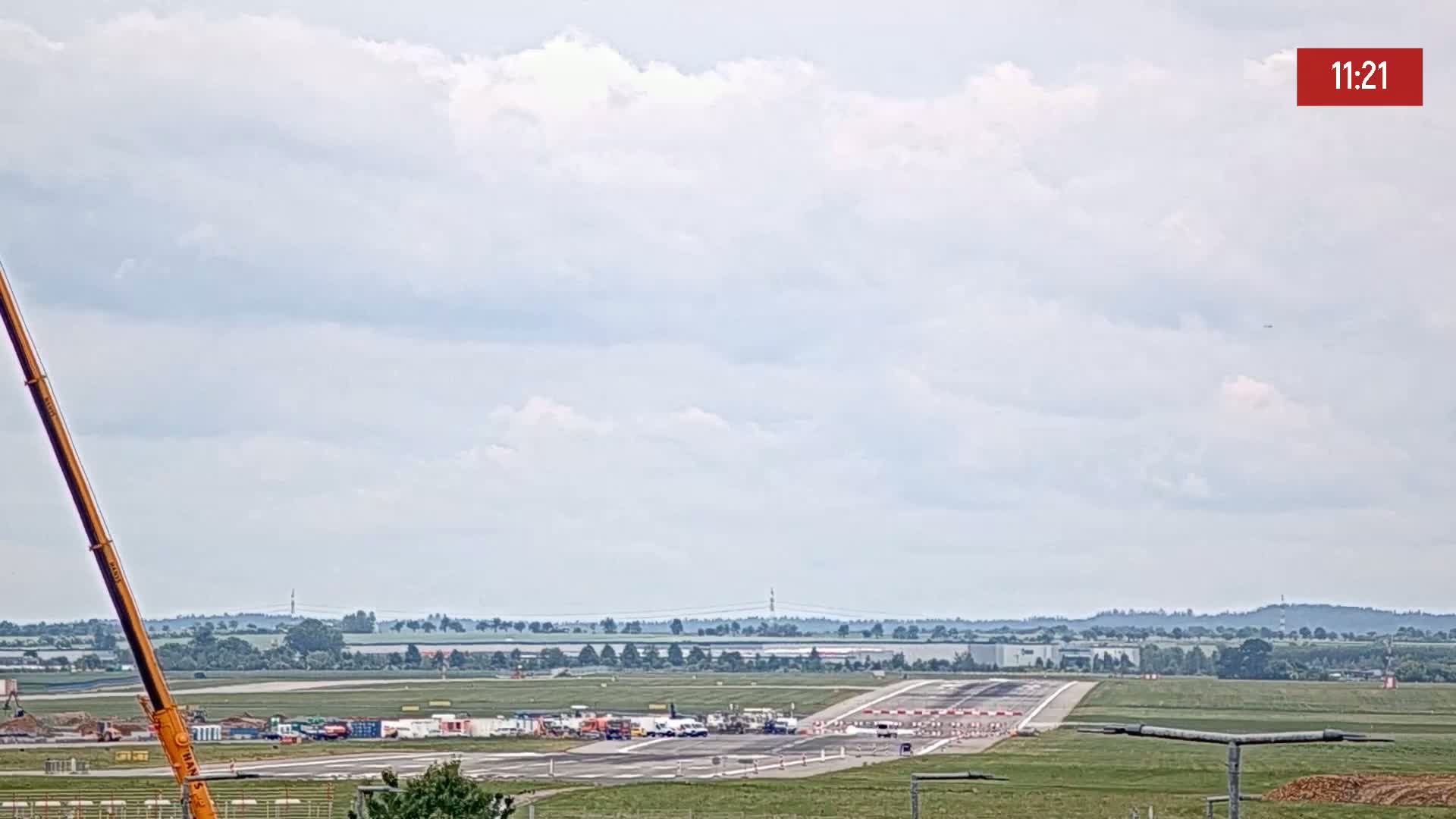 Under a partly cloudy sky, a long crane is visible to the left of an airport runway where several vehicles and construction equipment are gathered.