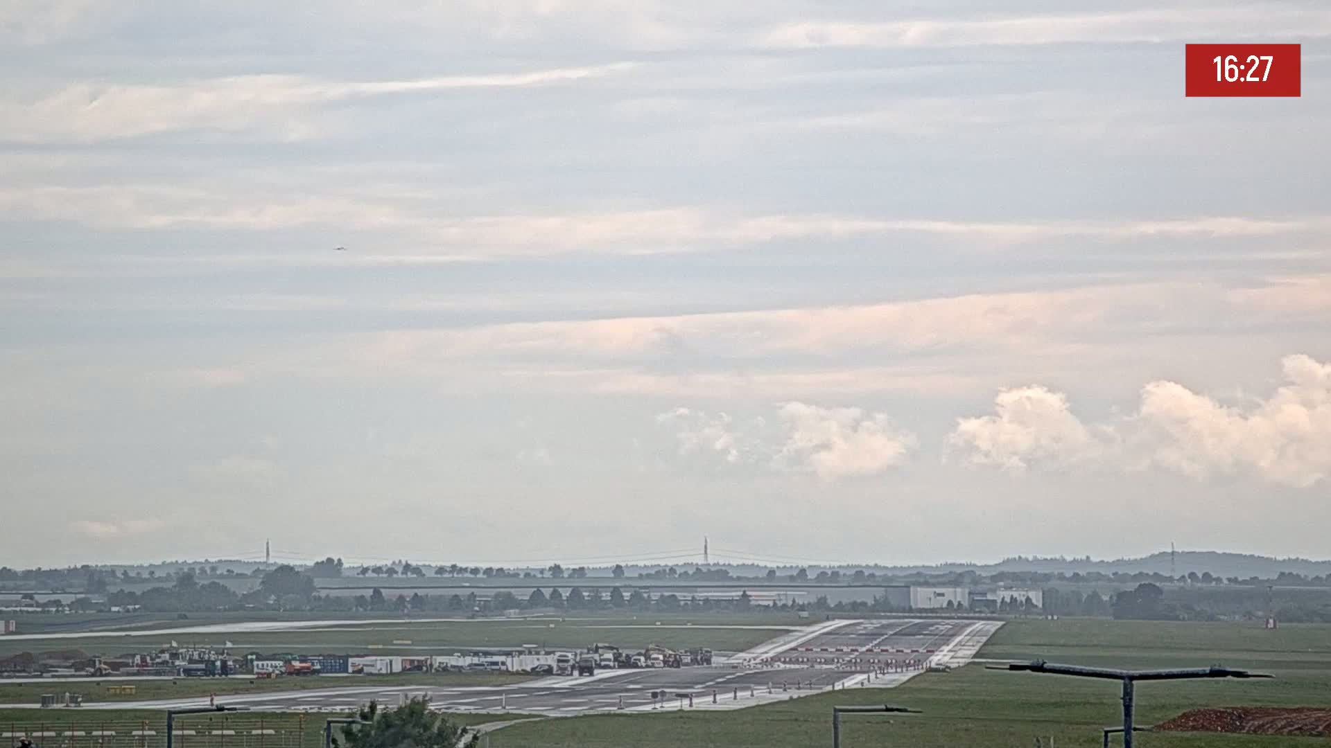 A long runway at an airport is visible under a mostly cloudy sky, with several vehicles parked near the end of the runway.