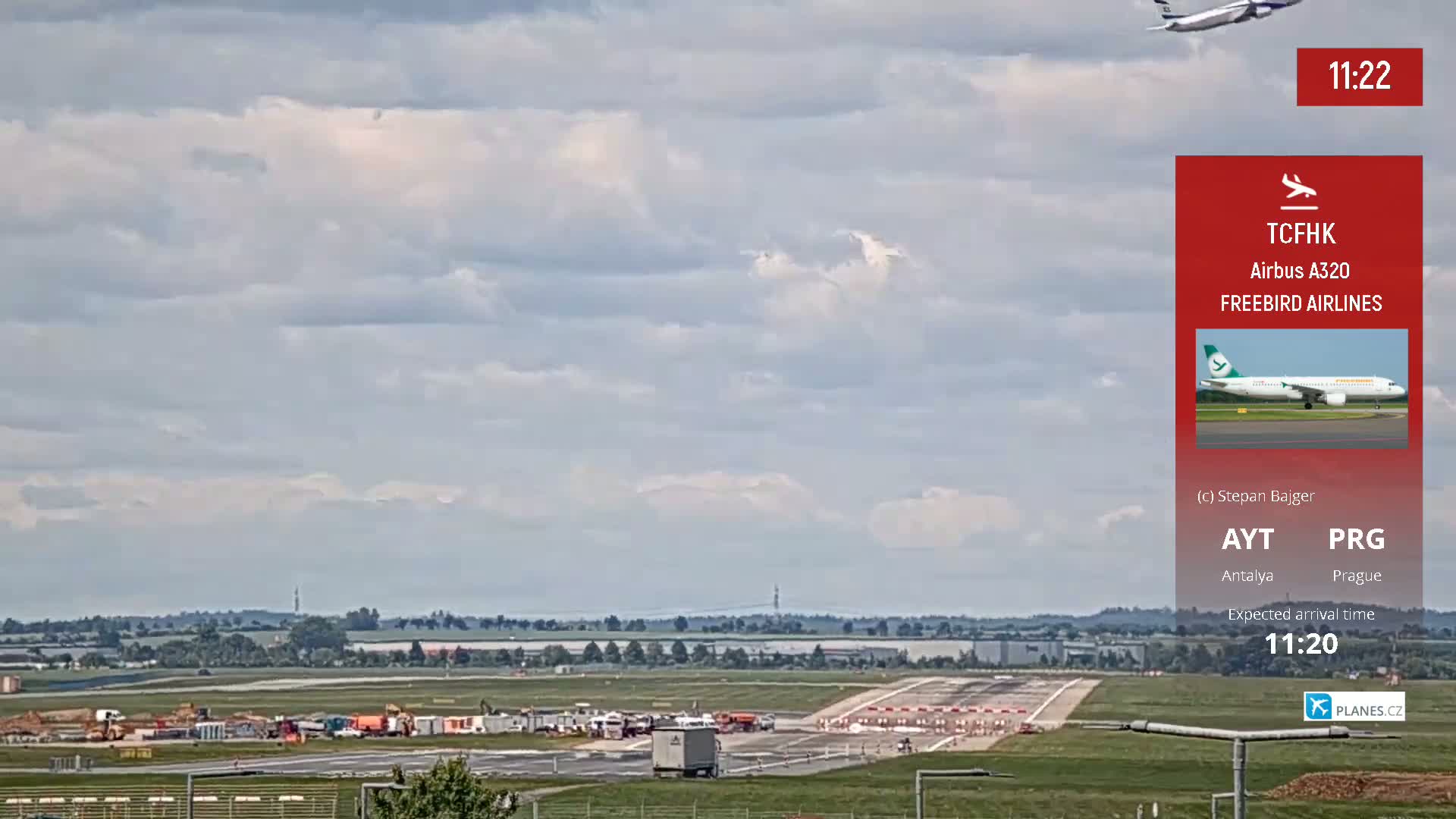 A mostly cloudy sky overlooks an airport runway with several vehicles and equipment visible, and a plane is seen approaching in the distance.