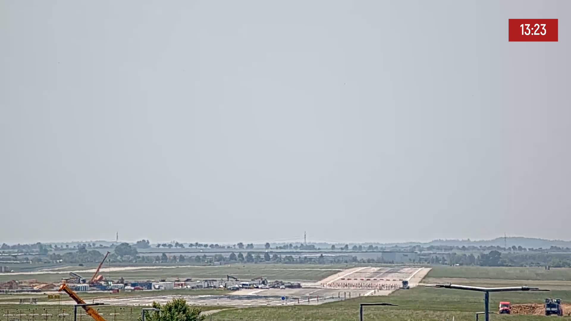 Under hazy skies, construction equipment is visible on a runway surrounded by flat, grassy terrain.