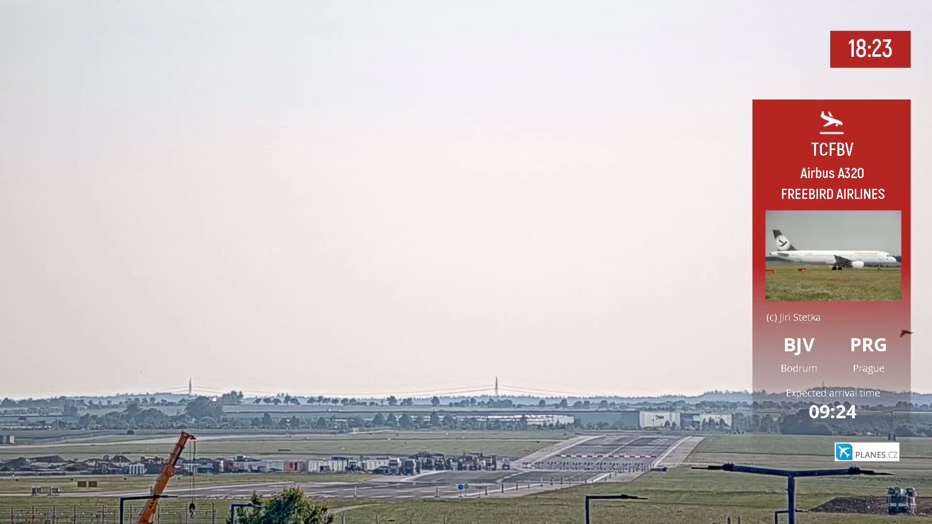A hazy, light-colored sky overlooks an airport runway with construction equipment and a Freebird Airlines Airbus A320 airplane visible in the distance.