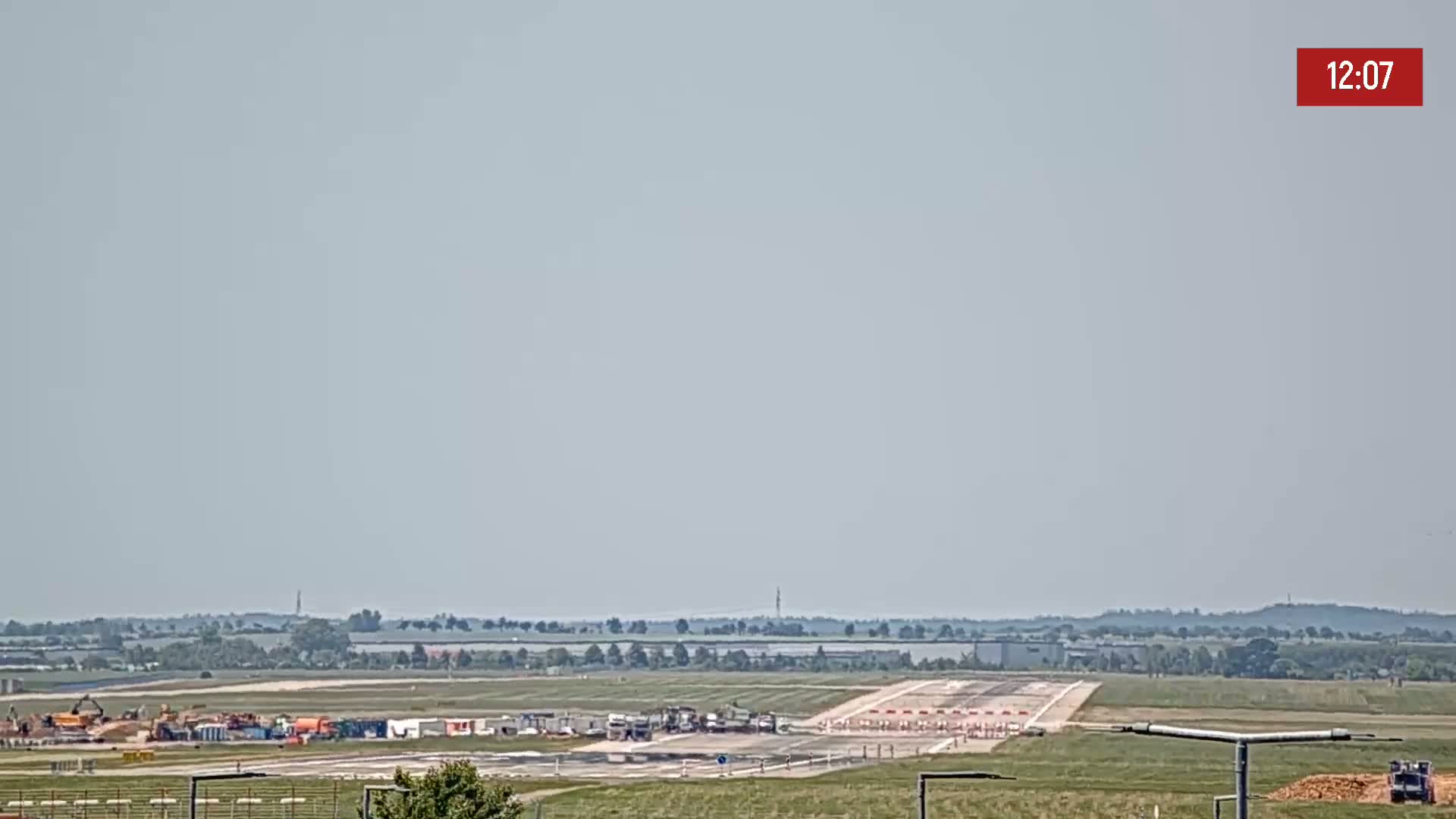 A long runway at an airport is visible under a hazy sky, with construction equipment and workers present on the airfield.