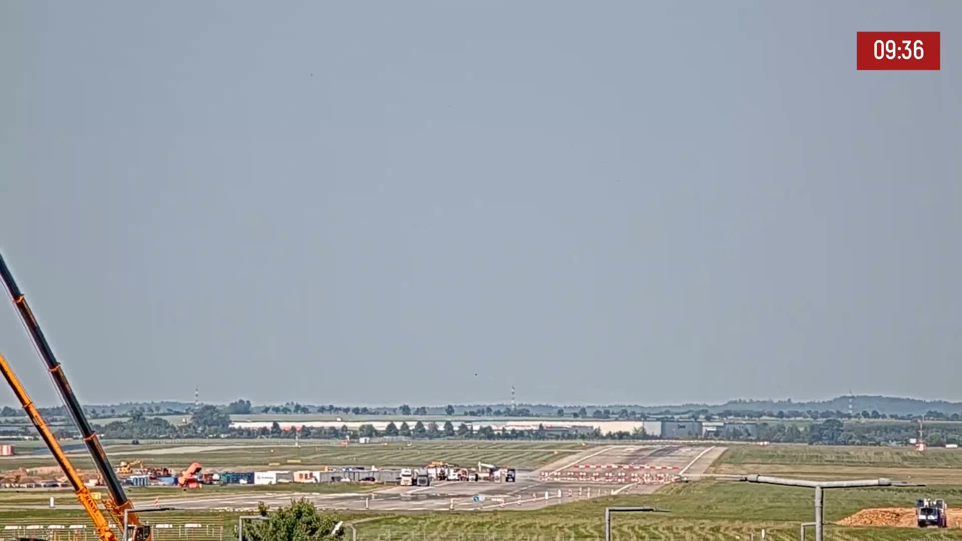Under a hazy sky, construction vehicles are visible on an airfield, with additional construction equipment and materials nearby.