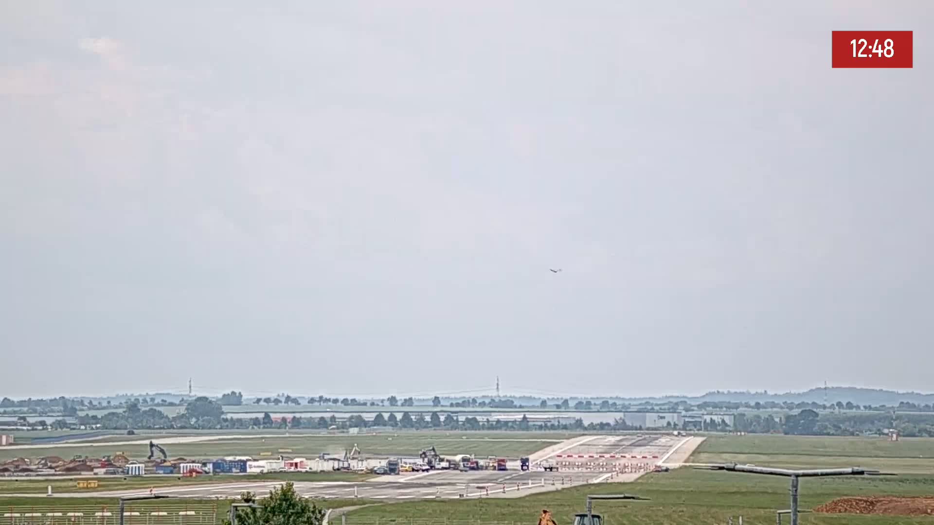 A long-range view of an airport runway under construction on an overcast day, with a single plane visible in the sky.