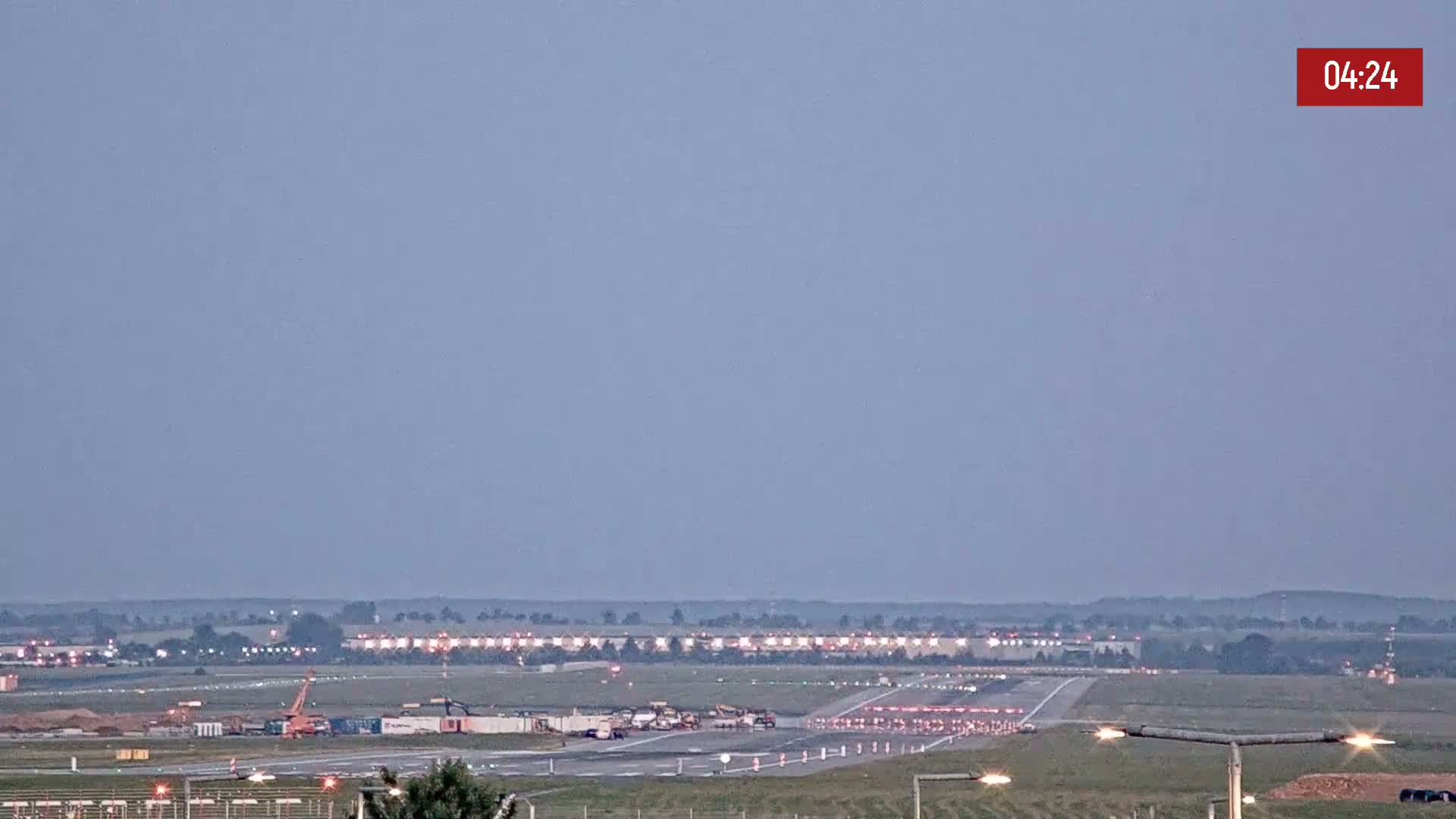 Under a pale, twilight sky, an airport runway is shown with construction equipment and vehicles present.