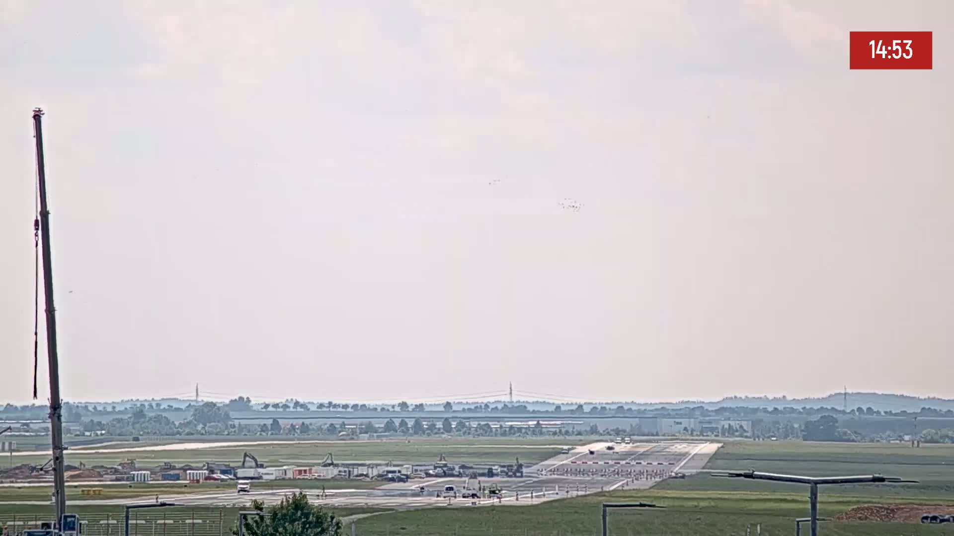 A long shot of an airport runway under hazy skies with construction equipment visible and a crane to the left, showing some vehicles on the tarmac.