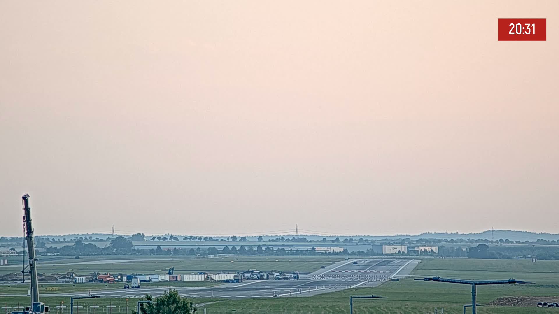 A hazy sunset illuminates a flat landscape featuring an airport runway with several vehicles and construction equipment nearby.