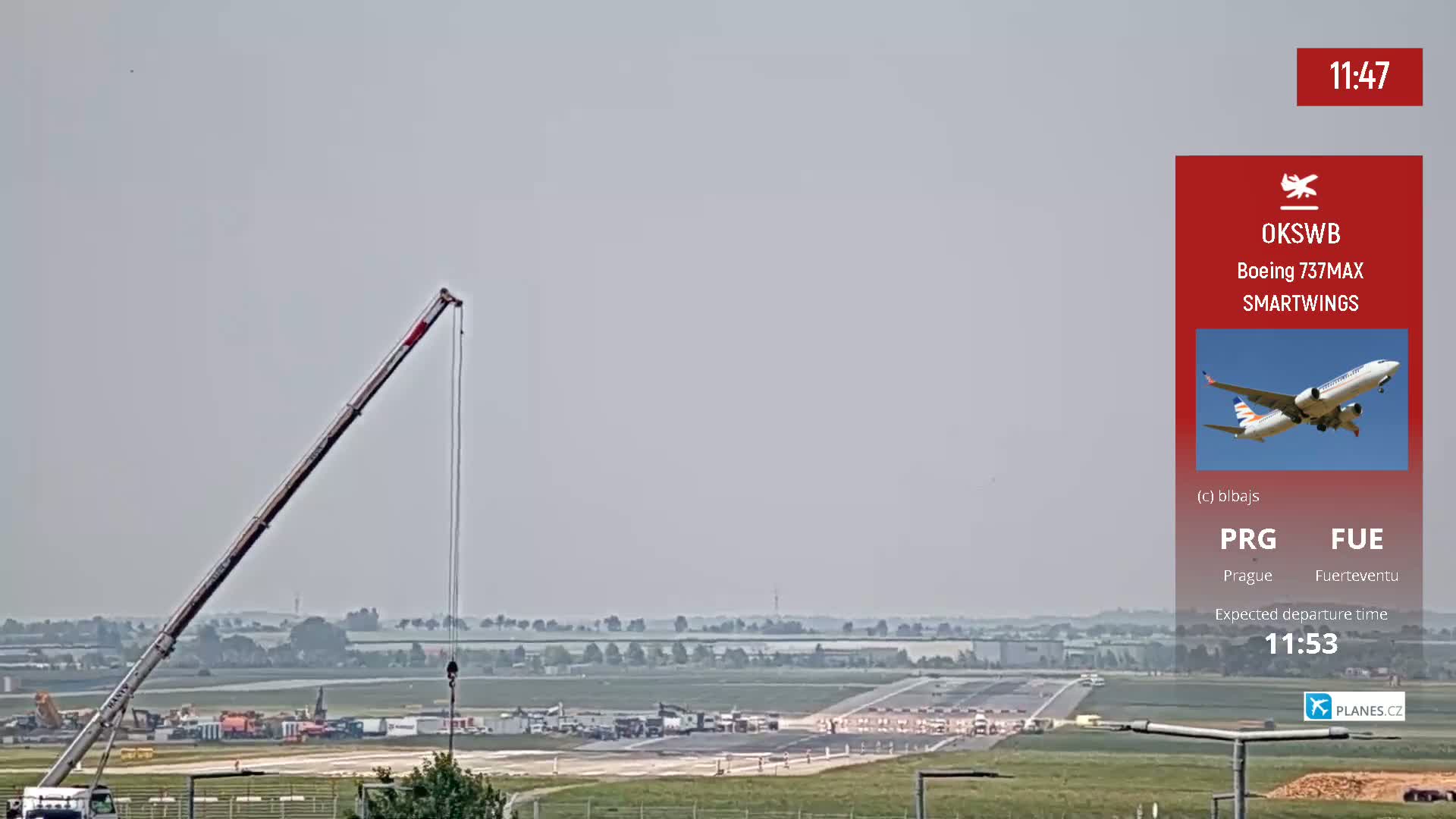 A large crane is visible on the left, overlooking an airport runway where various vehicles are parked and a plane is taking off under hazy sky conditions.