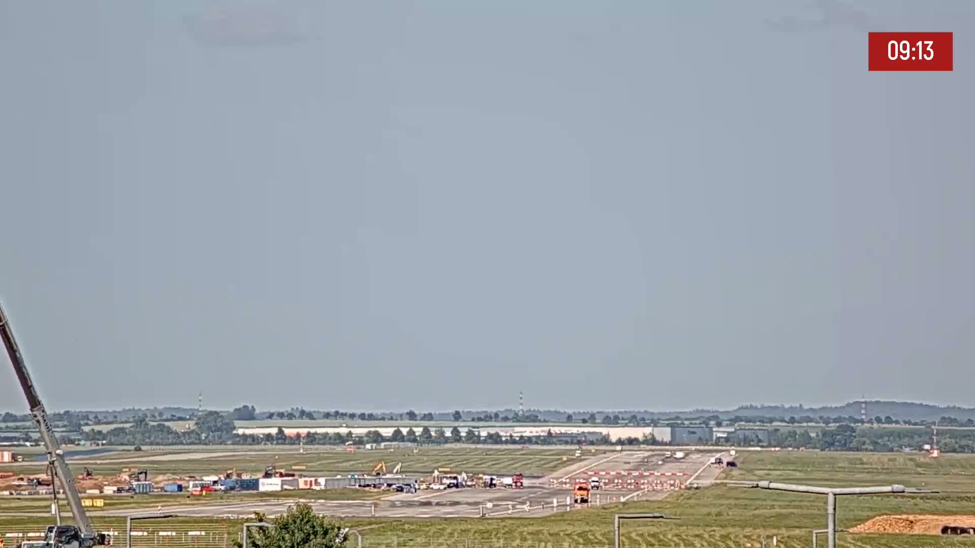 Under a hazy sky, construction equipment is visible on an airport runway, with flat, green land extending into the distance.