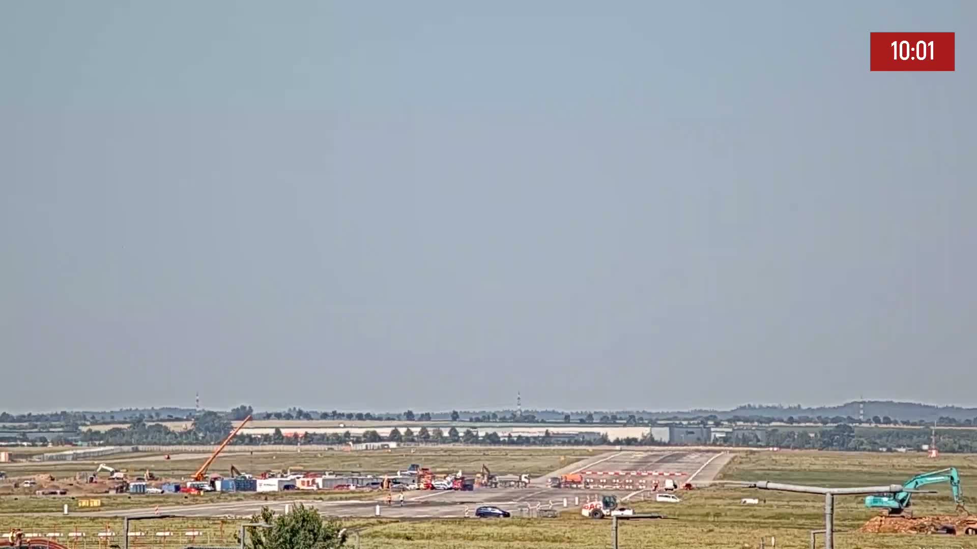 Under a hazy sky, construction equipment and vehicles are visible on a runway surrounded by flat land.