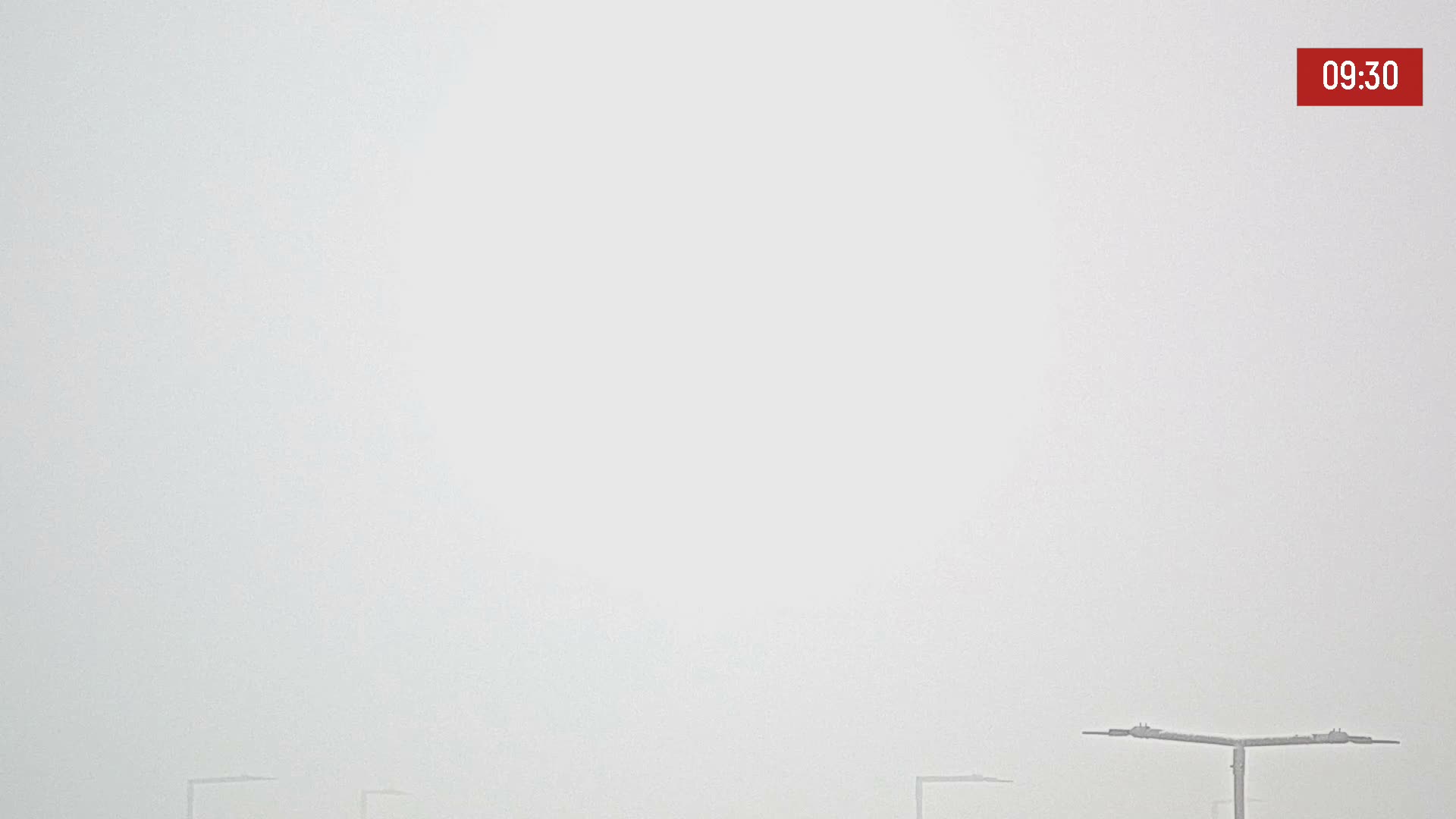 An outdoor scene is enveloped in extremely dense fog or whiteout conditions, revealing only the faint, dark outlines of structures barely visible at the bottom.