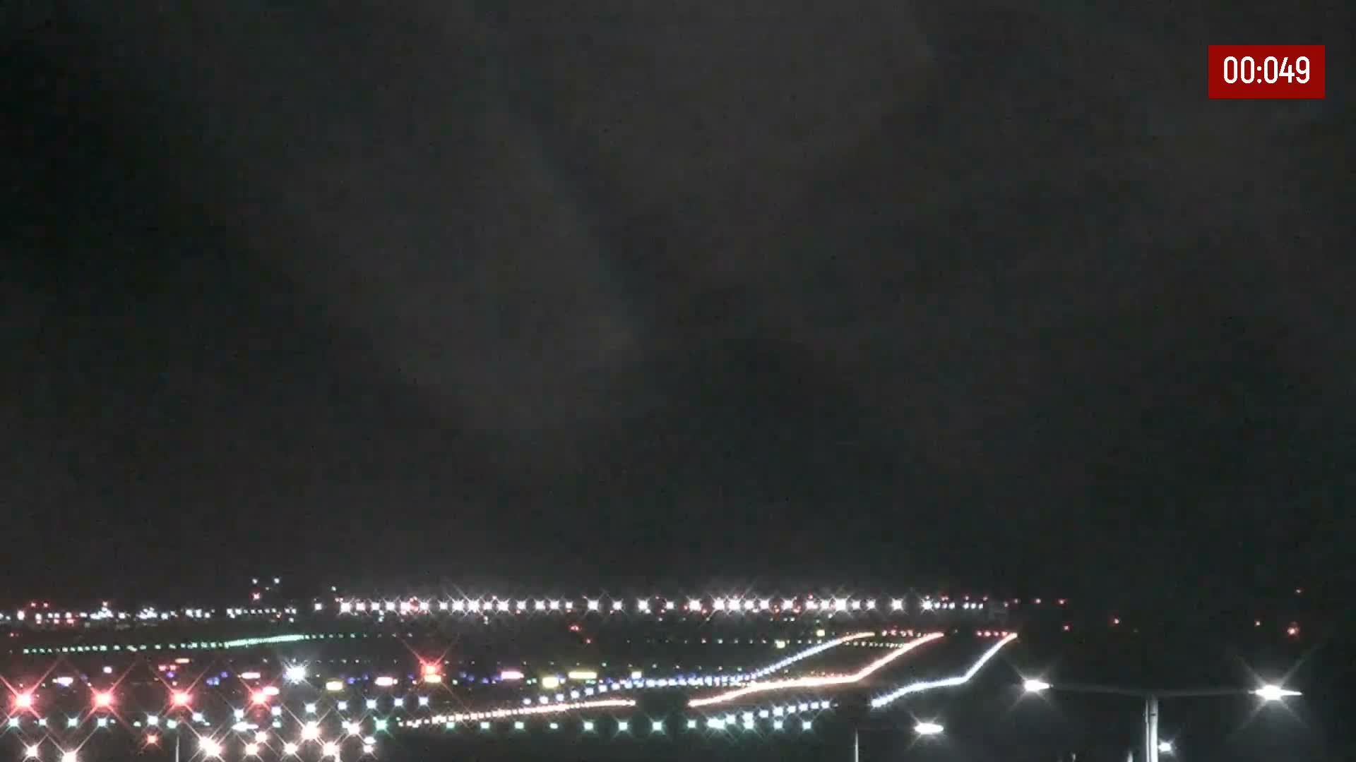 An overcast night sky hangs over a brightly illuminated airport runway and taxiway system, with numerous sparkling white, red, and colored lights dotting the ground.