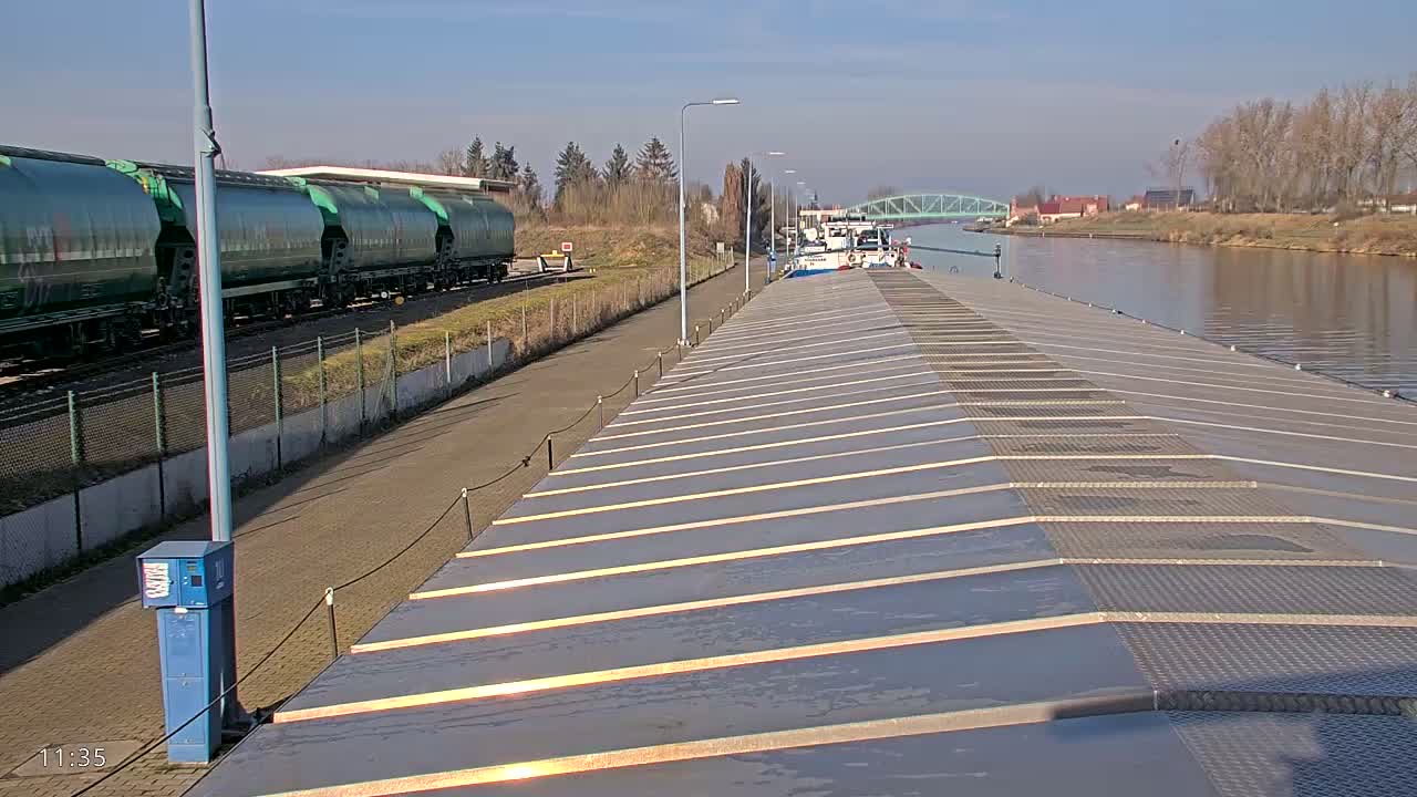 A barge is docked at a waterway under a sunny sky, next to a train of green tanker cars.