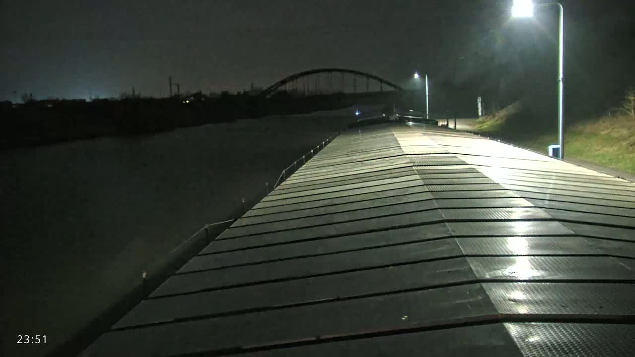 A long, dark, flat surface, possibly a barge or dock, stretches toward a lit bridge at night over calm water.