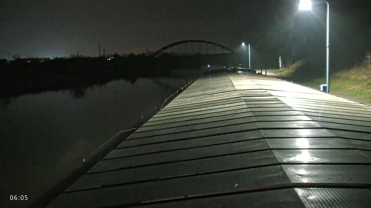 At night, a long, flat, metallic surface extends toward a distant arch bridge over a dark waterway, illuminated by several streetlights.