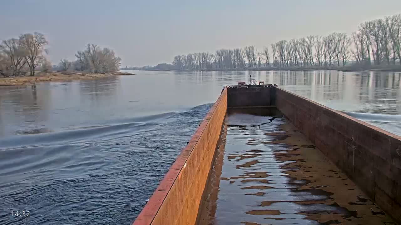 A rusty barge moves along a calm river under a clear sky, past bare winter trees lining the banks.