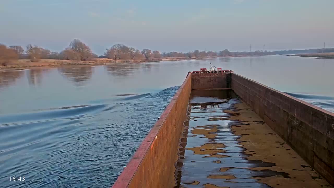 A rusty barge travels down a calm river under a hazy sky, leaving a wake in its path.