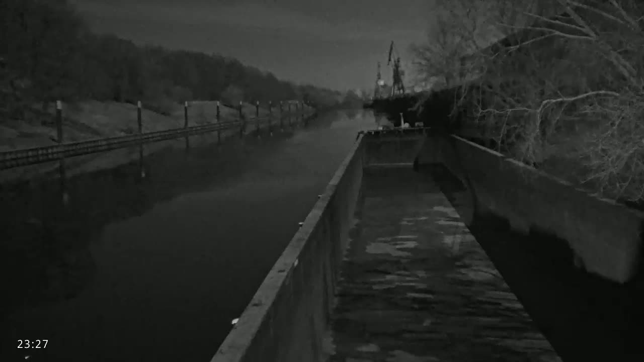 A nighttime view from a barge on a calm, dark river shows a bank with trees and a distant industrial area.