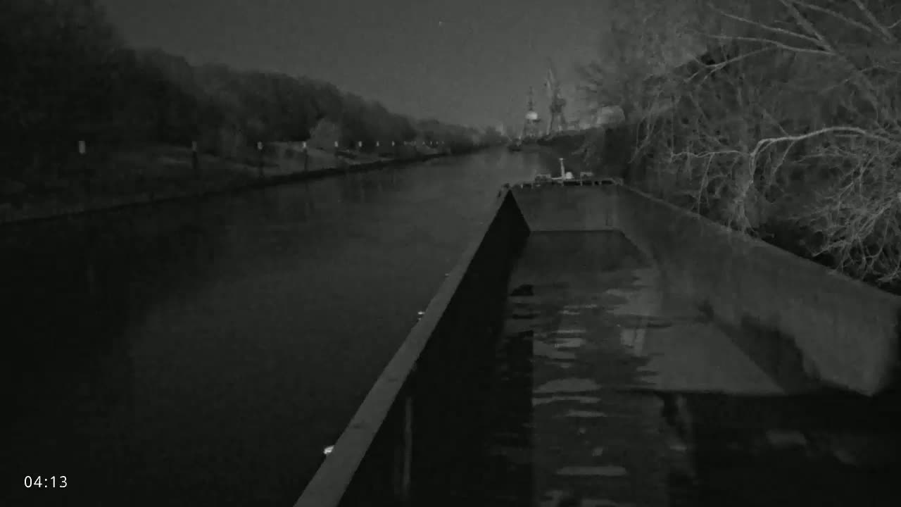 A barge travels down a dark canal at night under a cloudy sky, passing trees and industrial structures.