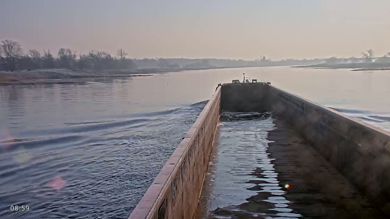 A barge moves along a calm river under a clear, light-grey sky, past a shoreline of bare trees.