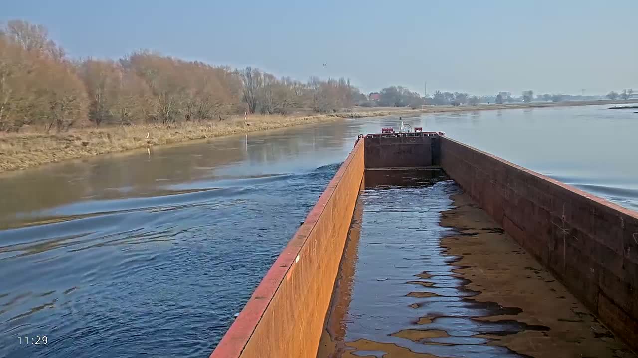 A rusty barge travels down a calm river on a clear, sunny day, past a shoreline of bare trees.