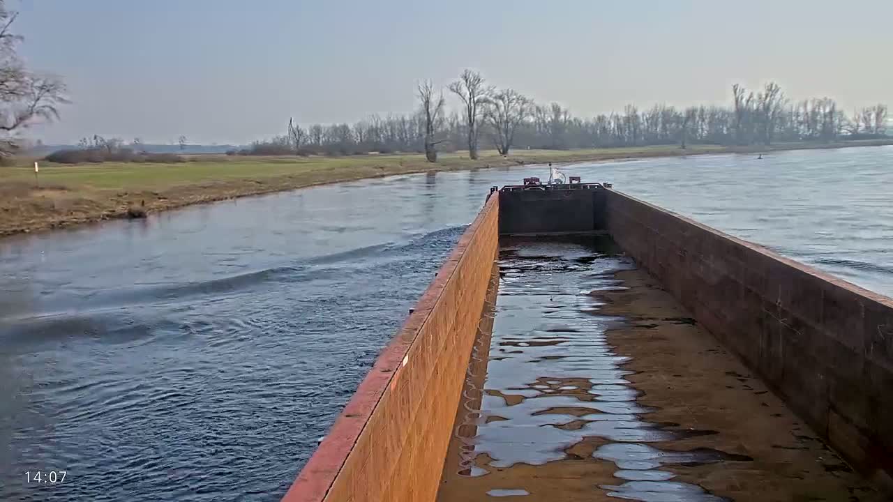 A barge travels down a river on a clear, sunny day, past a flat, grassy shoreline lined with leafless trees.