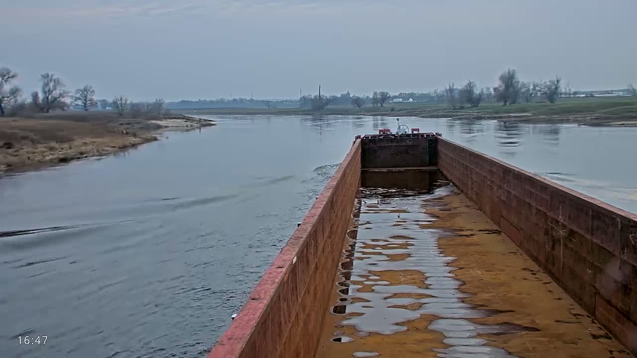 A rusty barge travels down a calm, gray river under an overcast sky, past a flat, treeless landscape.