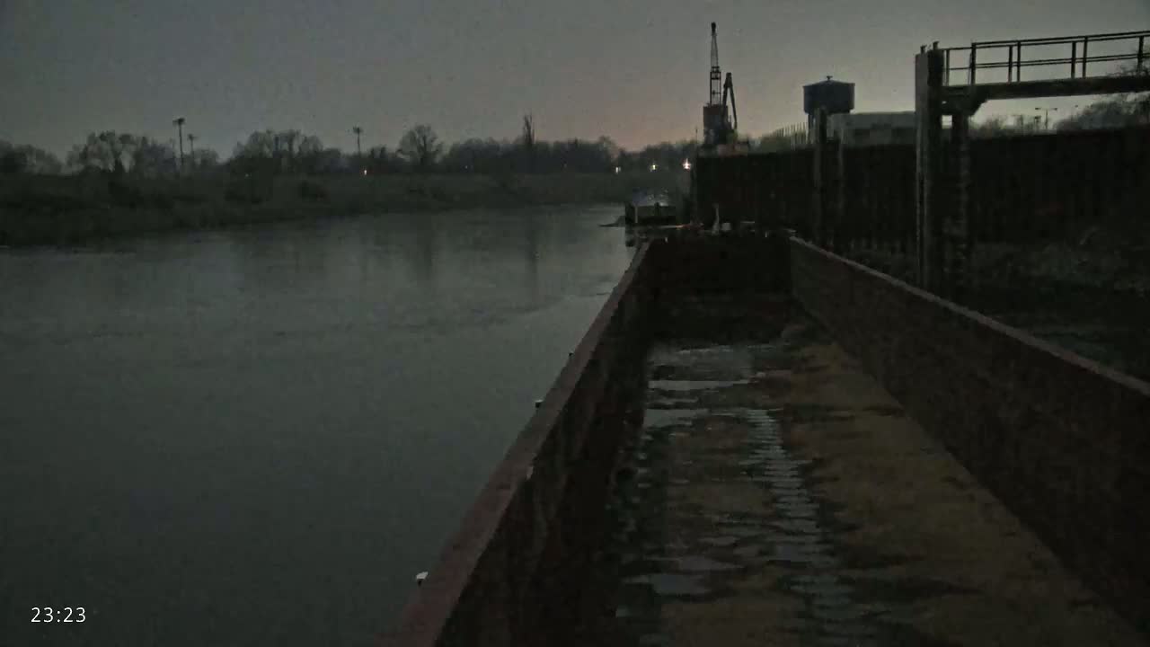 A long barge sits on dark, still water under a gray, overcast sky, next to a lock and a crane.