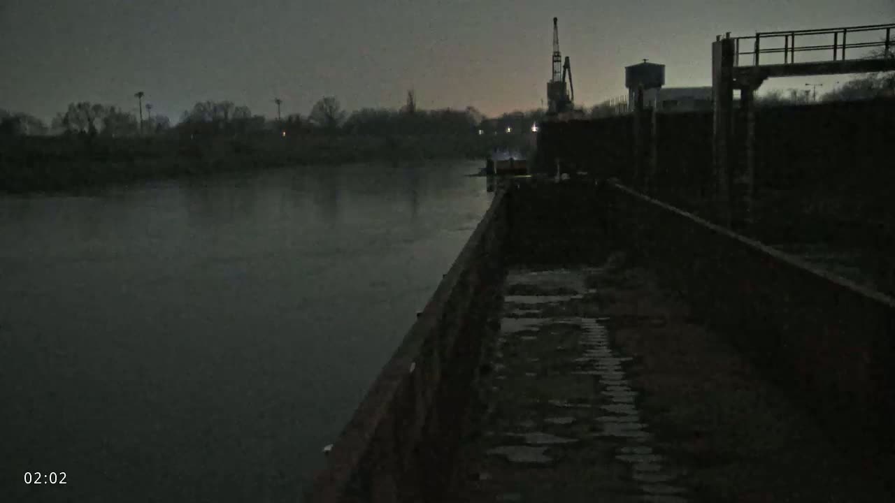 A dark, overcast night scene shows a barge alongside a calm river under a dim sky, with a crane and industrial structures visible in the background.