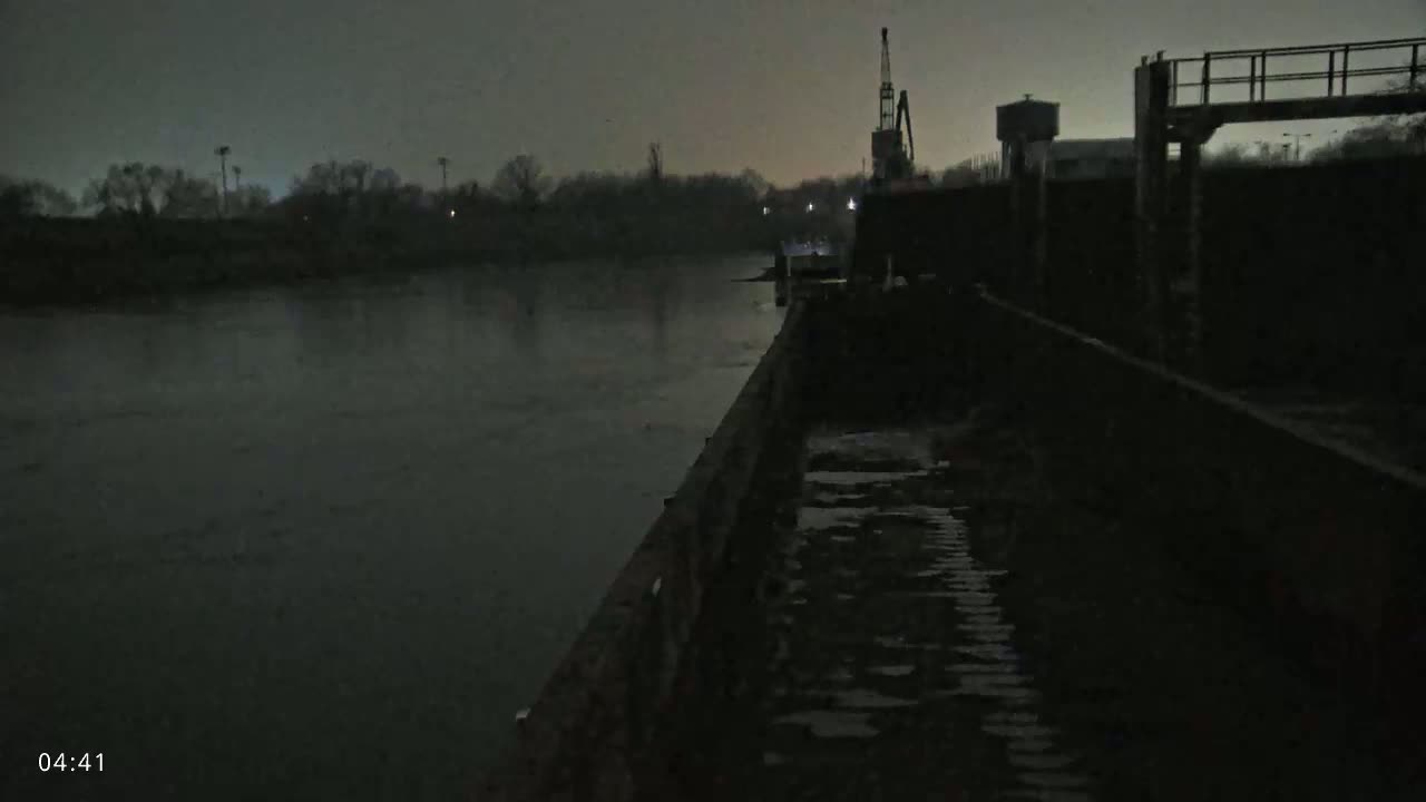 A dark, overcast night shows a river with a barge and industrial structures along the bank.
