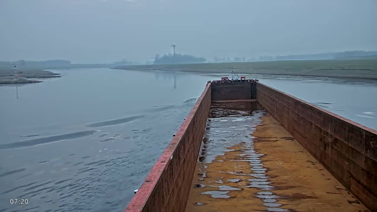 A rusty barge travels down a calm, foggy river toward a distant shoreline.