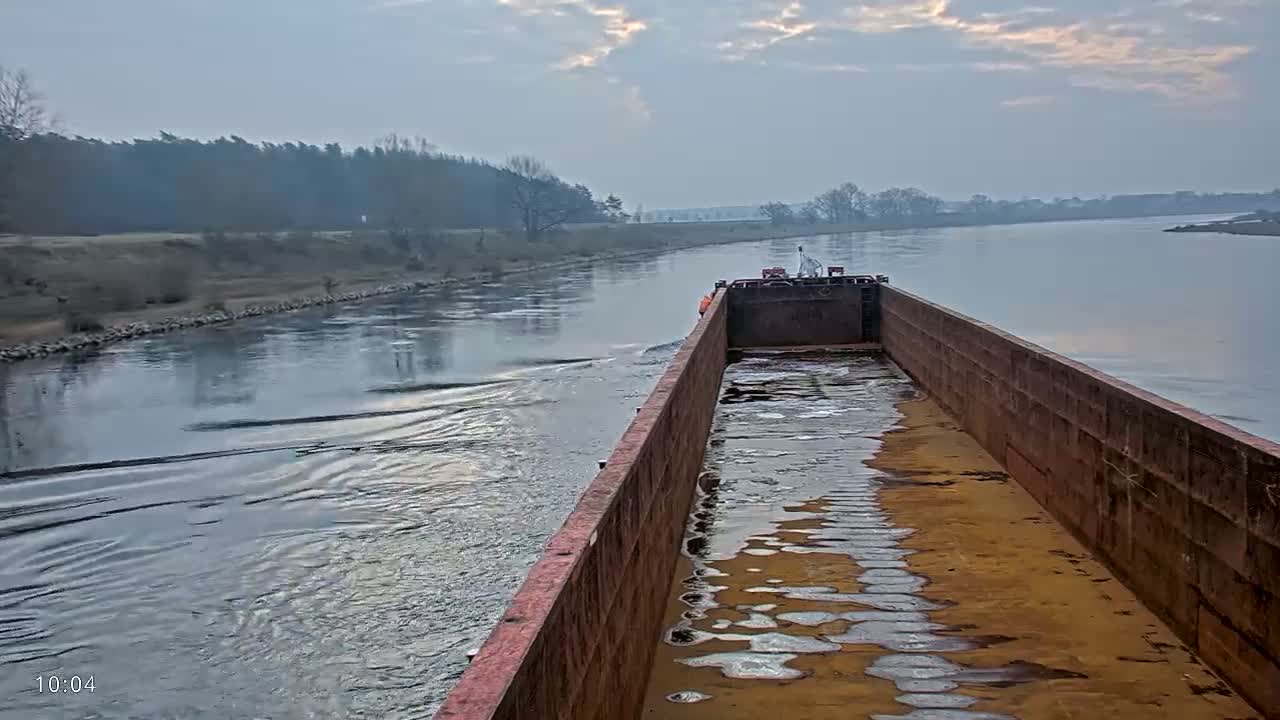 A barge travels down a calm river under a cloudy sky, with a shoreline of trees and low vegetation visible in the distance.