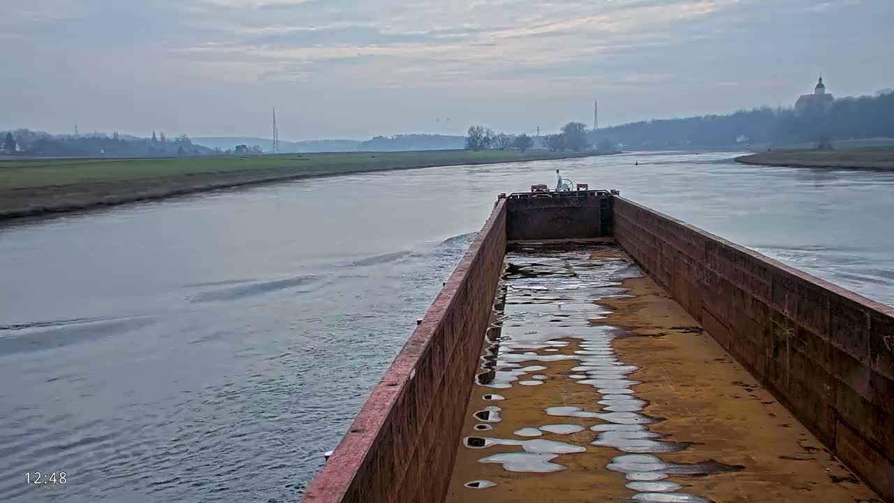 A barge travels down a calm river on an overcast day, with low-lying land and trees visible on either side.
