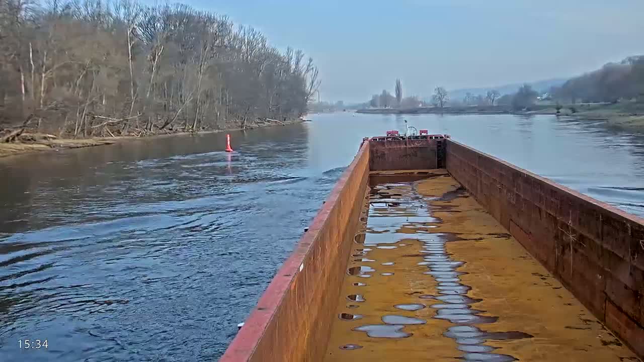 A rusty barge travels down a calm river past a treeline on a clear day.