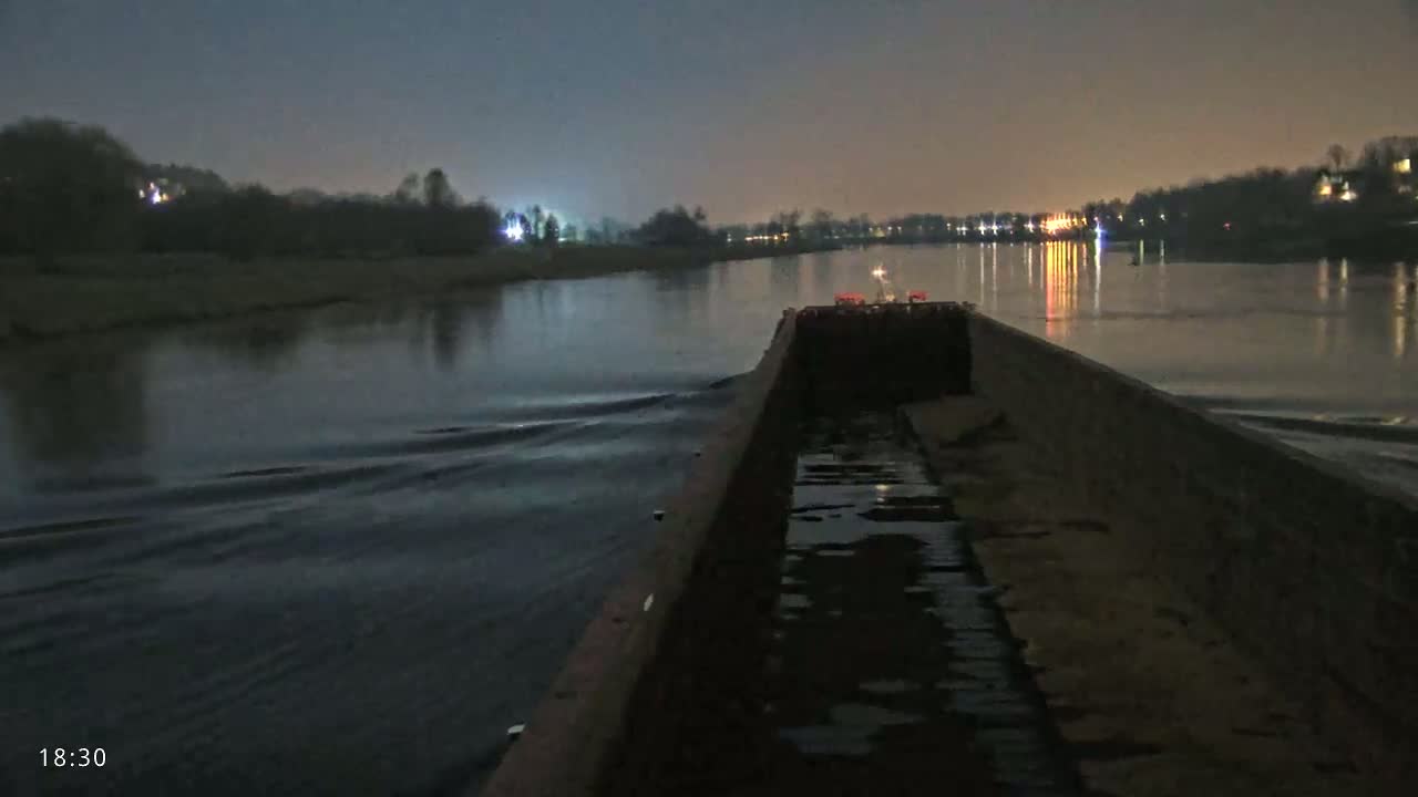 A barge travels on a dark river at night, under a mostly clear sky, past faintly lit shorelines.