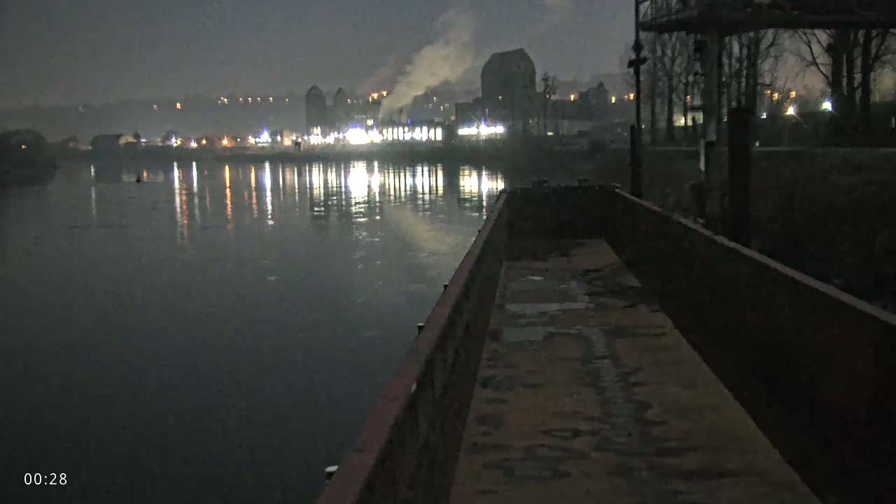 A nighttime view from a barge shows a calm river reflecting lights from buildings and smoke on the opposite bank under a hazy sky.