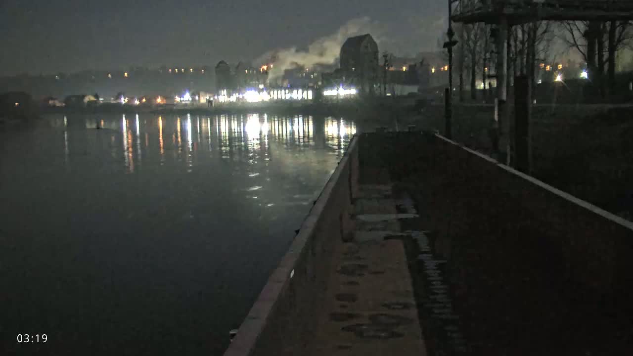 A nighttime view from a barge shows a dark, calm river reflecting the lights of a distant industrial area under a clear, dark sky.