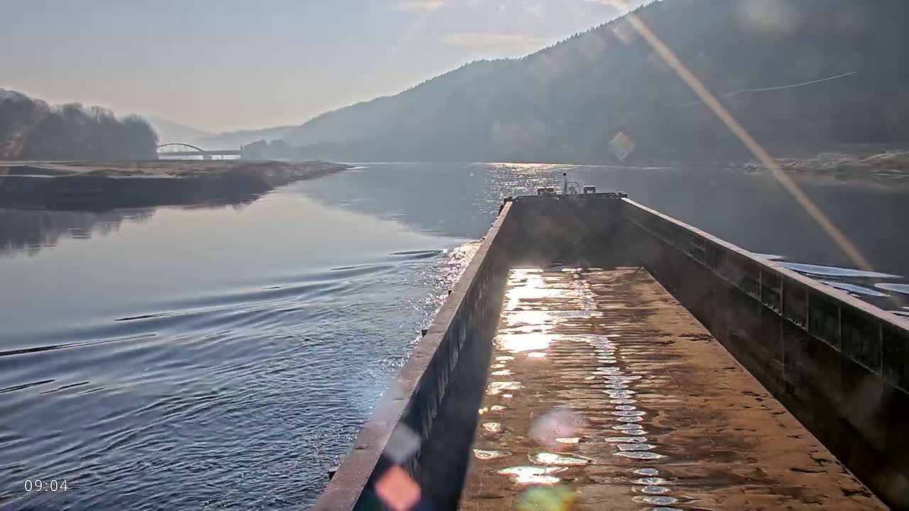 A barge travels down a river on a sunny, slightly hazy day, with forested hills visible in the distance.