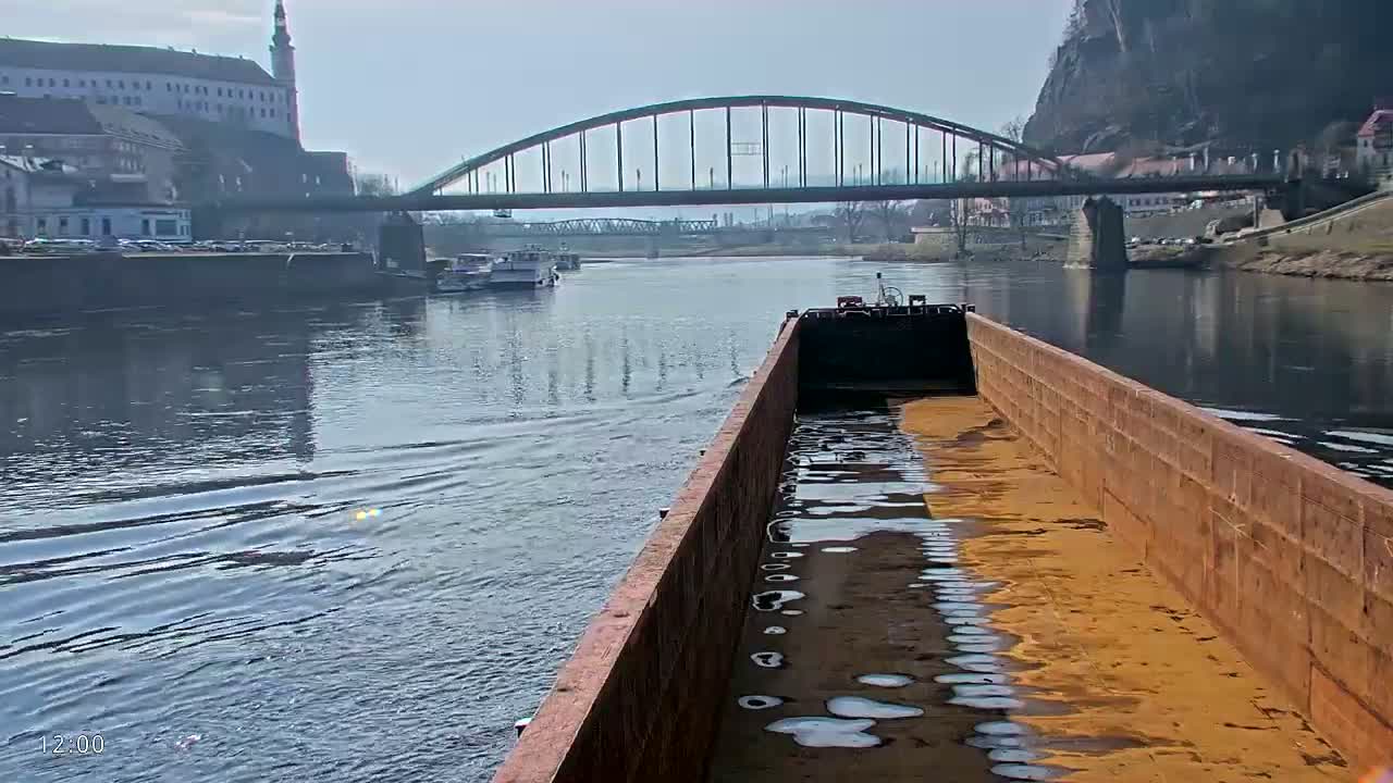A barge carrying a tan substance travels down a river on a partly sunny day, past a bridge and a castle.