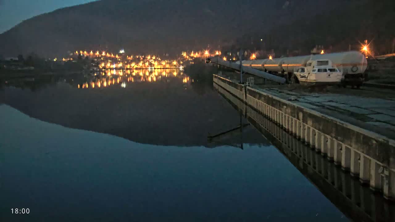 A long tanker truck and a small boat are parked at a dock at night next to a calm body of water reflecting the lights of a town on the opposite shore, under a dark sky.